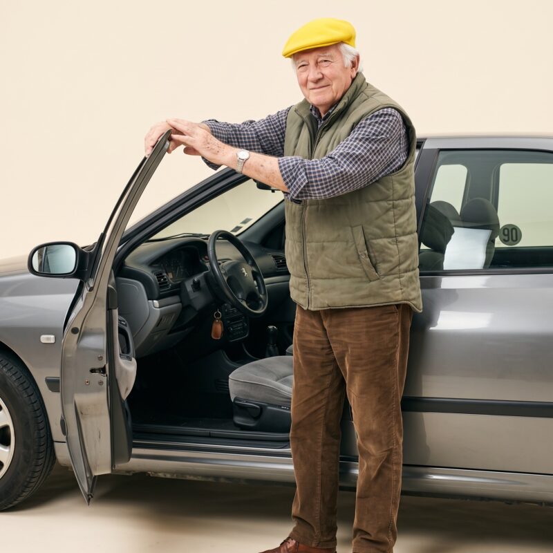 Portrait studio : retraité à casquette jaune debout devant sa berline des années 90, portière ouverte