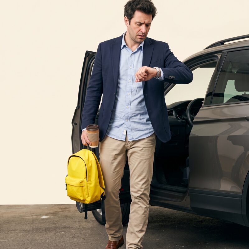 Portrait studio : parent pressé regardant sa montre devant son SUV portière ouverte, cartable jaune à la main