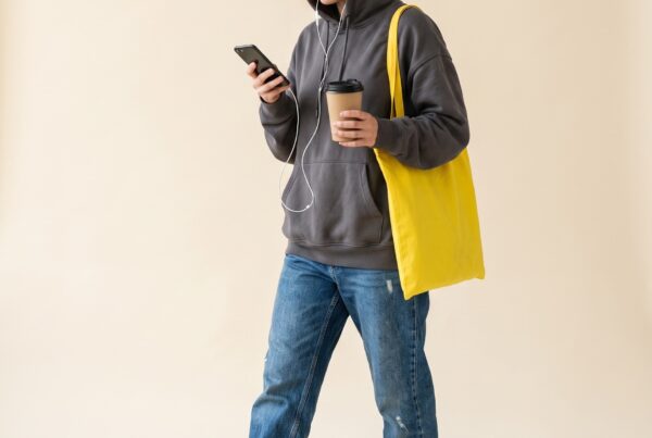 Portrait studio : piétonne capuchée absorbée par son téléphone, café à la main, tote bag jaune, un pied sur la chaussée